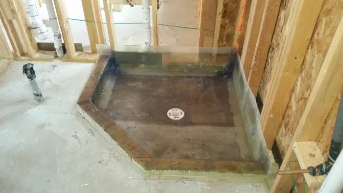 Angled-corner shower pan waterproofed across the floor and out over the curb, drain flange set in the sloped basin
